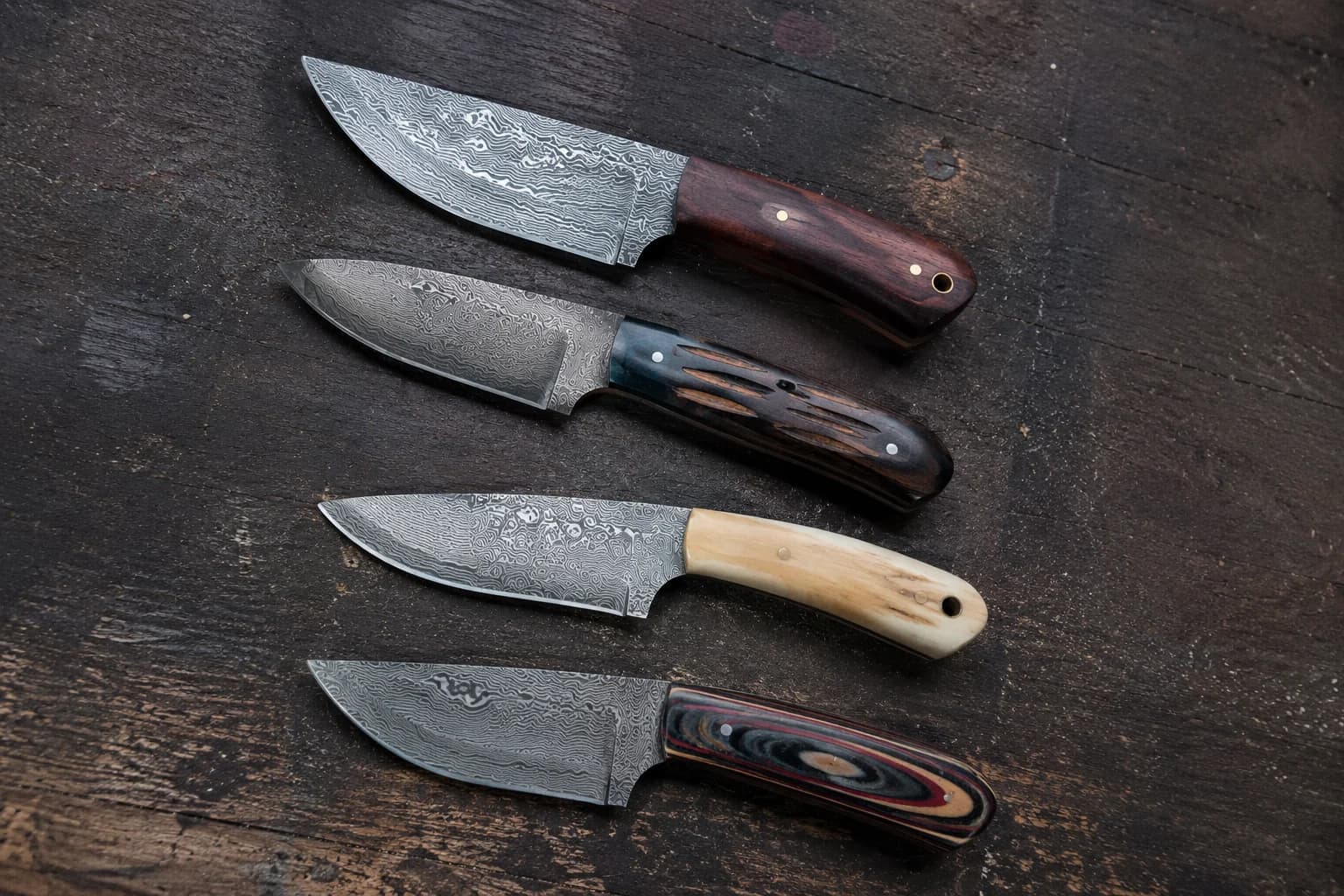 Four finished Damascus knives laid out on dark timber, each with a different handle material